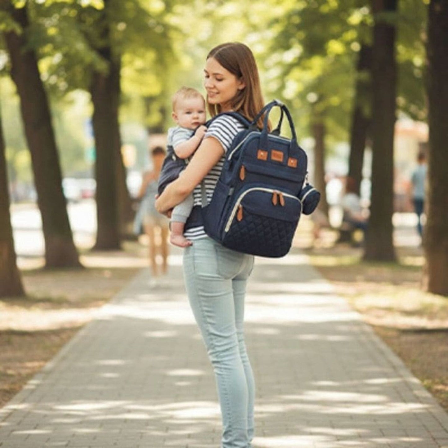 sac à dos à langer avec lit intégré Nomade porté par une maman tenant son bébé lors d’une promenade en extérieur 