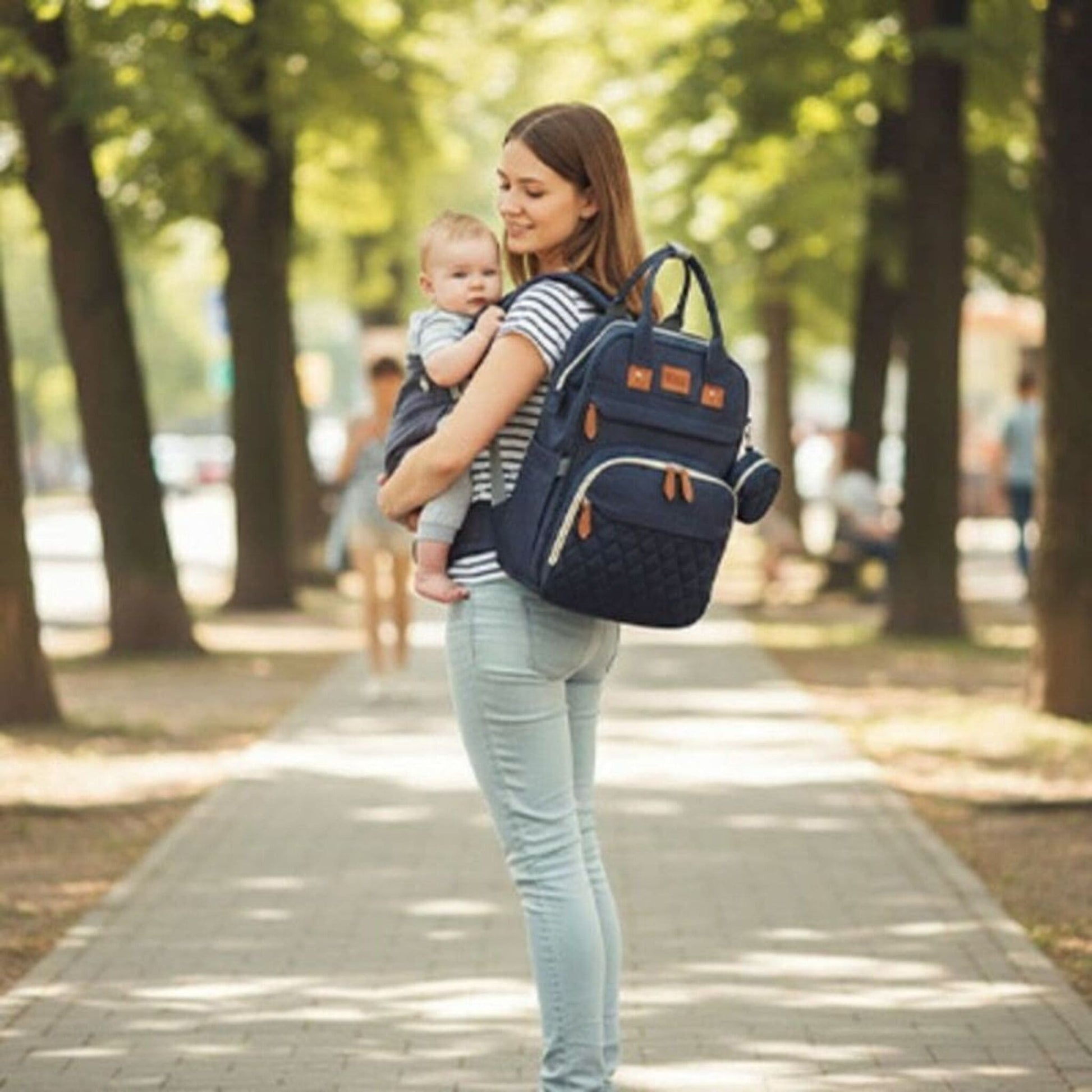 sac à dos à langer avec lit intégré Nomade porté par une maman tenant son bébé lors d’une promenade en extérieur 